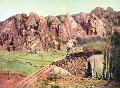 In a photo that was long ago tinted for effect, a Short Line 
to Cripple Creek train chugs over one of the line’s 31 timber 
railroad bridges as it passes through Cathedral Park in 1904. The Short Line, built by Irving Howbert as part of a
fierce railroad competition for Cripple Creek gold, evolved into Gold Camp Road.